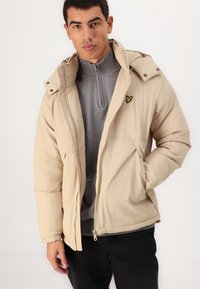 Beige puffjakke med hætte, lavet af letvægtsstof. Har lynlås lukning, forlommer og et lille gult logo på brystet.