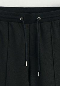 Ceinture élastique noire avec cordon de serrage ajustable, dotée d'aglets argentés. Tissu doux avec détail plissé à l'avant et ouverture pour poche latérale.