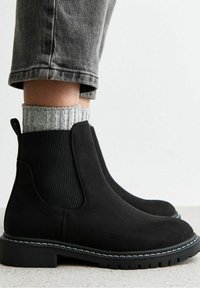 Bottines Chelsea en daim noir avec des panneaux élastiques sur les côtés et une languette, complétées par une chaussette en maille côtelée grise texturée et une semelle robuste.