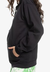 Sweat à capuche noir avec une poche kangourou, des poignets et une ceinture côtelés. Texture lisse, coupe décontractée, présenté de profil sur un fond neutre.