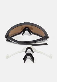 Zonnebril met een zwart frame en bruine lenzen, met verwisselbare neusstukjes in zwart en wit, die een sportief ontwerp uitstralen.