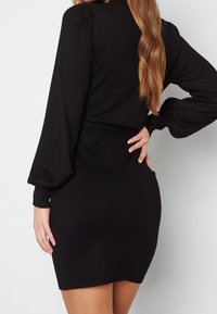 Robe noire ajustée avec manches longues bouffantes et taille cintrée. Matière douce avec une texture lisse, se terminant juste au-dessus du genou.