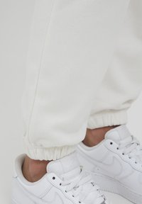 Weiße Sweatpants mit elastischen Bündchen, aus weichem Stoff gefertigt. Kombiniert mit weißen Sneakers, die ein dezentes Logo und Strukturen an der Seite aufweisen.