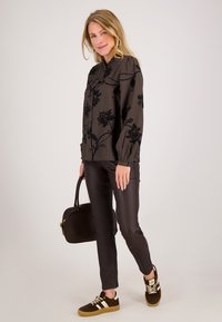 Donkere bloemenblouse met opgelegde mouwen, gecombineerd met strakke donkere broek en bruine suède sneakers met witte strepen. Model draagt een bruine handtas.