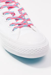 Białe płócienne trampki z gumową podeszwą, z różowymi i niebieskimi plecionymi sznurowadłami oraz strukturalnym czubkiem. Zbliżenie na przód buta.