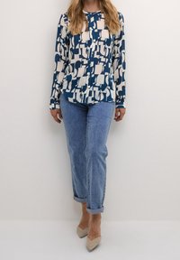 Langärmliges blaues Hemd mit einem cremefarbenen abstrakten Muster, mit Knöpfen auf der Vorderseite. Kombiniert mit hellblauen hochgekrempelten Jeans und beigen Heels.