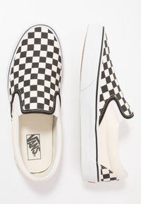 Zapatillas de lona a rayas en blanco y negro, tipo slip-on, con suelas de goma blancas y costuras en contraste, que cuentan con un logo de Vans en la plantilla.