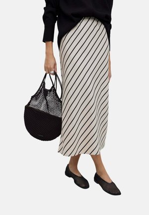 Mujer con top negro, falda larga beige con rayas diagonales negras, bailarinas negras de malla y sosteniendo un bolso negro de red.