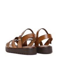Sandali in pelle marrone con cinturini intrecciati, fibbie regolabili alla caviglia e suola scura e texturizzata di media altezza. Presentano un design piatto per il massimo comfort.