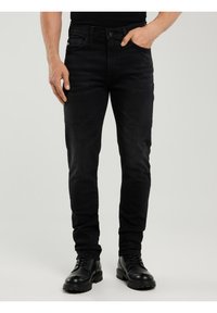 Bărbat purtând blugi negri slim-fit și cizme negre din piele, stând cu o mână în buzunar pe un fundal simplu.