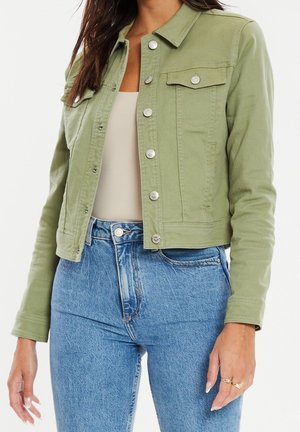 Kvinde iført lysegrøn knapjakke over beige top og højtaljede blå jeans, stående med afslappede arme.