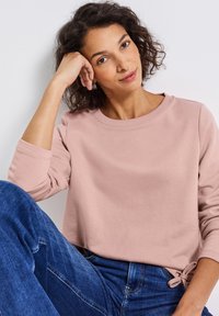 Blushfärgad sweatshirt med rund hals och långa ärmar, tillsammans med blå denimjeans. Sweatshirten har en sidoknytning.