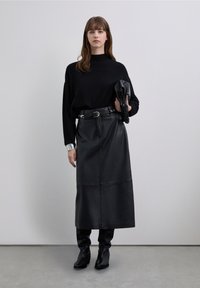 Pulover negru tricotat cu guler înalt, asortat cu o fustă midi neagră din piele, cu textură netedă și linii clare, accesorizată cu o curea.
