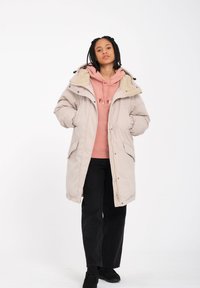 Lys beige parka med hætte, blødt indre for, to frontlommer og lynlås lukning over en pink hoodie og sorte bukser.