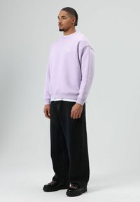 Sweat-shirt lilas avec poignets côtelés, associé à un jean noir ample et des chaussures noires épaisses. Design minimaliste sans motifs ni logos visibles.