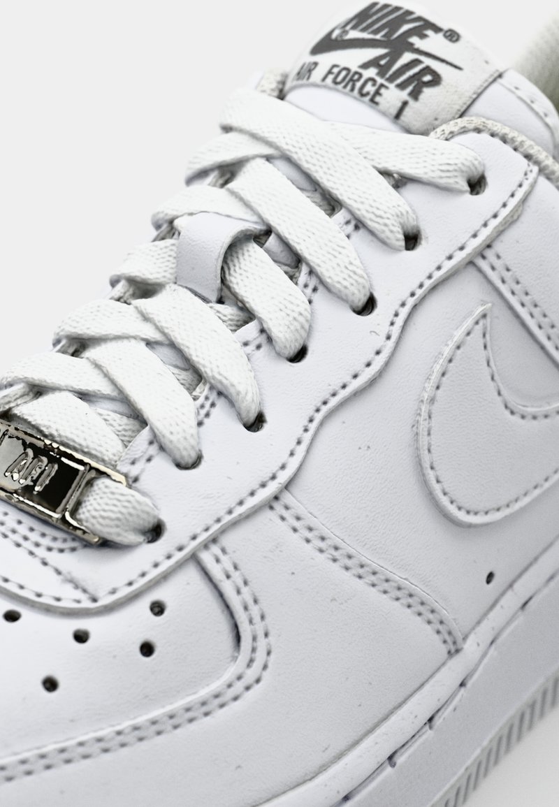 Sneaker Nike Air Force 1 en cuir blanc avec des lacets plats blancs, une étiquette métallique pour les lacets et des coutures visibles le long des côtés et de la semelle.