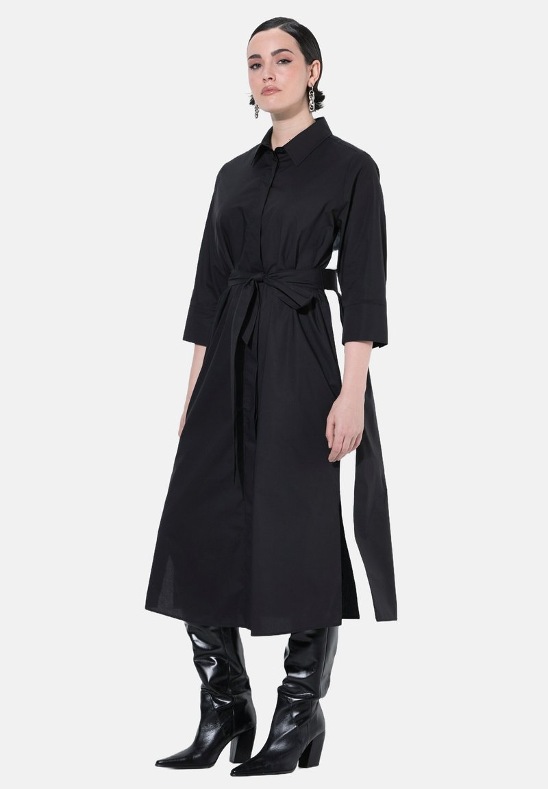 Vestito nero a camicia con colletto, maniche lunghe e vita legata, realizzato in tessuto liscio. Abbinato a stivali neri lucidi fino al ginocchio.