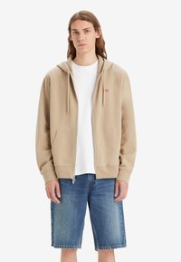Beige rits-trui met een voorzak en een capuchon met koord, gedragen over een wit T-shirt, gecombineerd met blauwe denim shorts.