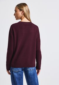 Pull en mailles bourgogne avec un col rond et des poignets côtelés, associé à un jean bleu. Texture simple sans motifs ni accents visibles.