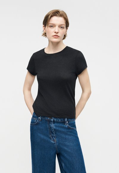 Νέα γυναίκα με κοντά μαλλιά, ντυμένη με μαύρο t-shirt και μπλε τζιν, στέκεται με τα χέρια στις τσέπες μπροστά σε απλό φόντο.