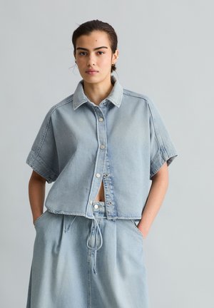 Junge Person trägt ein locker sitzendes, hellblaues Denim-Hemd mit kurzen Ärmeln und Knöpfen sowie dazu passende, hochtaillierte Denim-Hosen und hat die Hände in den Taschen.