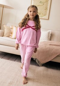 Ensemble de vêtements d'intérieur en polaire rose comprenant un haut à manches longues orné d'un grand nœud rouge et un pantalon assorti. Texture douce et coupe décontractée.