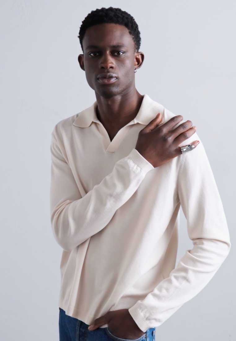 Polo à manches longues, couleur beige clair, avec col, poignets côtelés et coupe décontractée, associé à un jean en denim foncé et un accessoire en forme d'anneau.