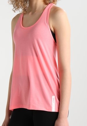 Femme portant un haut rose sans manches ample par-dessus un soutien-gorge de sport noir, associé à un pantalon noir, sur un fond clair uni.