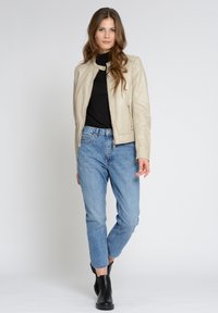 Beige Lederjacke mit Reißverschluss, getragen über einem schwarzen Rollkragenpullover. Kombiniert mit hellblauen high-waisted Jeans und schwarzen Stiefeletten.