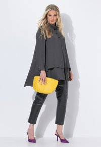Blonde woman in loose dark gray tunic, black leather pants, purple heels, holding textured yellow clutch bag, posing against white wall.