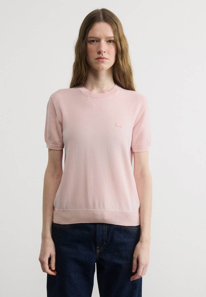 Νεαρή γυναίκα με μακριά μαλλιά φοράει ένα ανοιχτό ροζ πουλόβερ με κοντά μανίκια και σκούρα μπλε τζιν, στέκεται μπροστά σε ένα απλό λευκό φόντο.