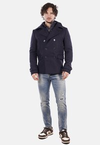 Cappotto doppiopetto navy in lana, con grandi bottoni, tasche laterali e una fodera interna trapuntata, abbinato a jeans strappati e sneakers.