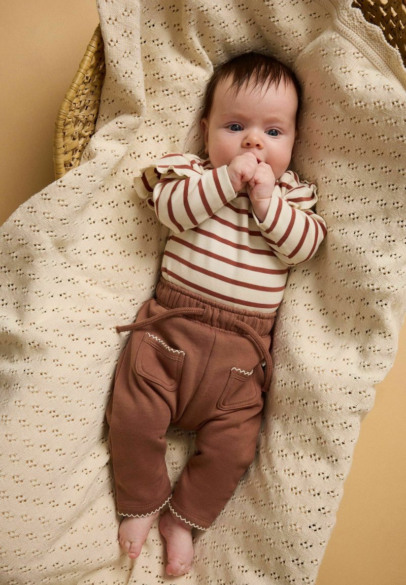 Bébé allongé sur une couverture tricotée crème dans un panier en osier, portant un t-shirt rayé à manches longues et un pantalon marron, les mains près de la bouche.