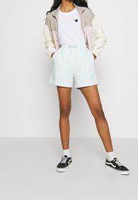 Modelka má na sobě barevně blokovanou bundu světle růžové, béžové a bílé barvy přes bílé tričko s černým srdcem, v kombinaci s mátově zelenými šortkami a černými teniskami.