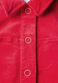 Röd kord skjorta med spetsig krage, två framfickor och rosa, runda knappar längs knäppningen. Mjuk textur med vertikala ribbor.