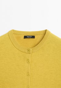 Žlutý pletený cardigan s kulatým výstřihem a čtyřmi knoflíky vpředu, uvnitř límce je viditelná značka Massimo Dutti.