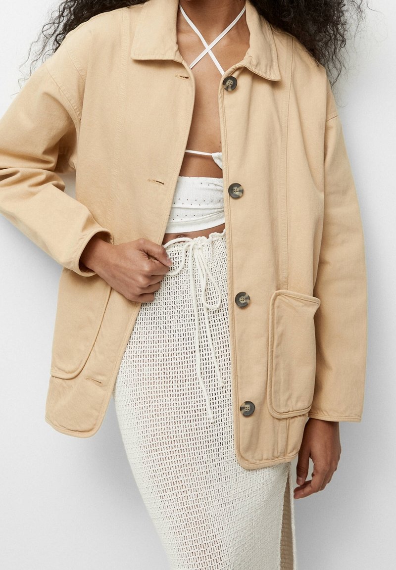 Vrouw in een beige knopenjasje over een witte gehaakte rok en een witte crop top met kruisbanden, hand die het jasje vasthoudt bij de voorkant.