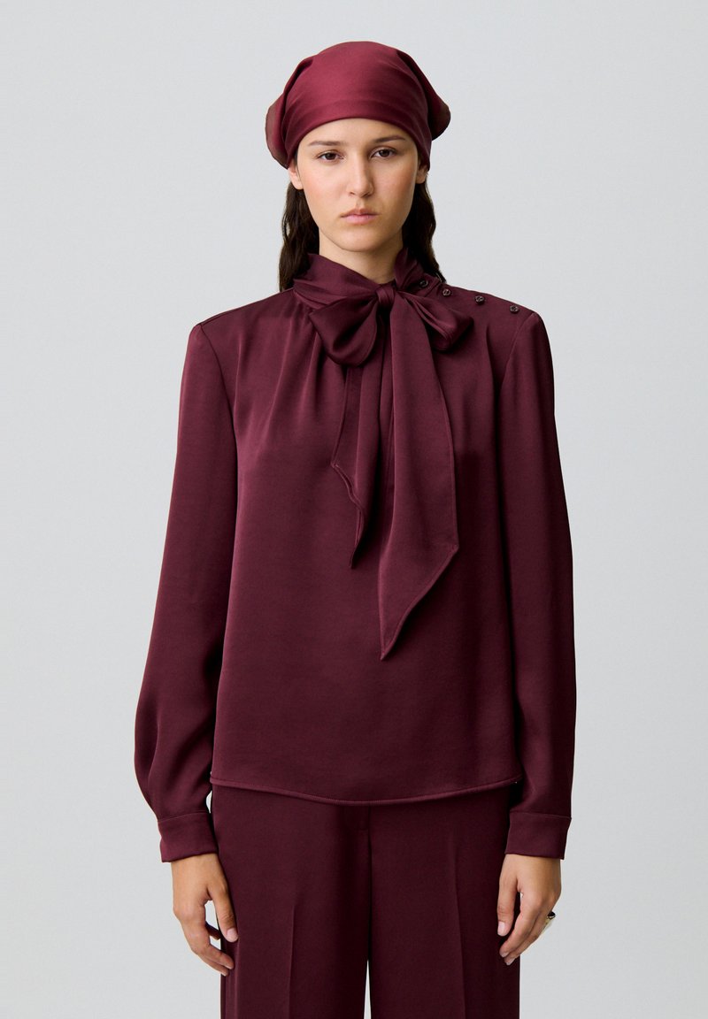 Blusa color granata con dettaglio di fiocco al collo, maniche lunghe e bottoni decorativi sulla spalla. Tessuto liscio con una sottile lucentezza.