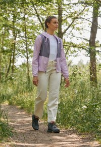 Giacca leggera di color viola chiaro e grigio con cappuccio, indossata sopra una camicia bianca e pantaloni beige. Scarpe da trekking nere. Circondata da vegetazione.