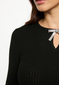 Top nero a coste con scollatura a chiave, impreziosito da un accento a fiocco di cristallo sul colletto. Il materiale sembra morbido e testurizzato.
