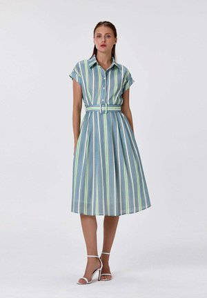 Femme portant une robe midi ceinturée à manches courtes avec col, à rayures verticales bleues, vertes et blanches, associée à des sandales blanches à talons.