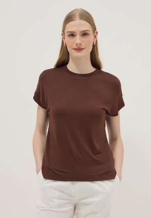 Mujer con el cabello largo y rubio, lleva pendientes de aro dorados, una camisa marrón de manga corta y pantalones blancos, de pie con las manos en los bolsillos.