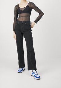 Persona vistiendo una blusa negra de manga larga transparente, pantalones rectos de tiro alto negros y zapatillas blancas con acentos azules, sobre un fondo liso.