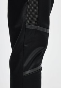 Pantalones deportivos negros con una textura suave, que cuentan con un acento negro brillante y un panel de malla con pequeños agujeros para transpirabilidad.