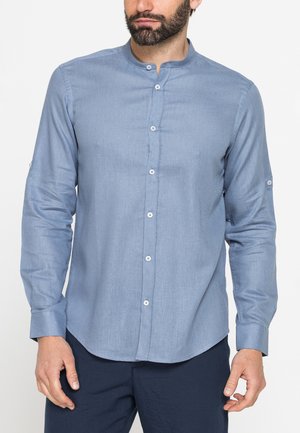 Uomo che indossa una camicia azzurro chiaro a manica lunga con collo alla mandarino e pantaloni scuri, in piedi contro uno sfondo semplice.