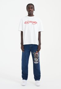 White short-sleeve t-shirt with red "Ed Hardy" logo, paired with blue jeans featuring colorful embroidered skull and serpent design. White sneakers.