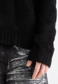 Pullover preto de malha com uma textura suave e felpuda e punhos canelados, combinado com calças escuras e texturizadas com padrões abstratos claros.