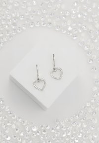 Silvriga hjärtformade örhängen med en glittrande kant och öglor som hänger, placerade på en vit kvadratisk pelare omgiven av kristaller.