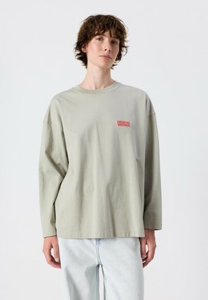 Junge Person mit kurzen braunen Haaren trägt ein lockeres beiges Langarmshirt mit dem Text "American Vintage" und hellblaue Jeans.