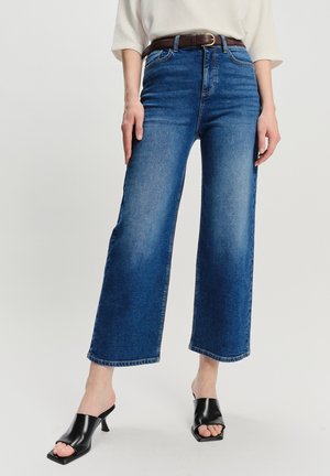 Donna che indossa jeans ampi a vita alta blu con una cintura marrone, top bianco e mule con tacco aperto neri, in piedi su uno sfondo bianco.